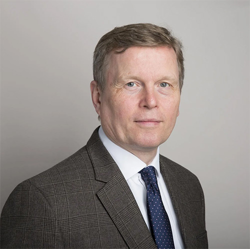 A man with gray hair wearing a gray blazer and dark blue tie looks at the camera.
