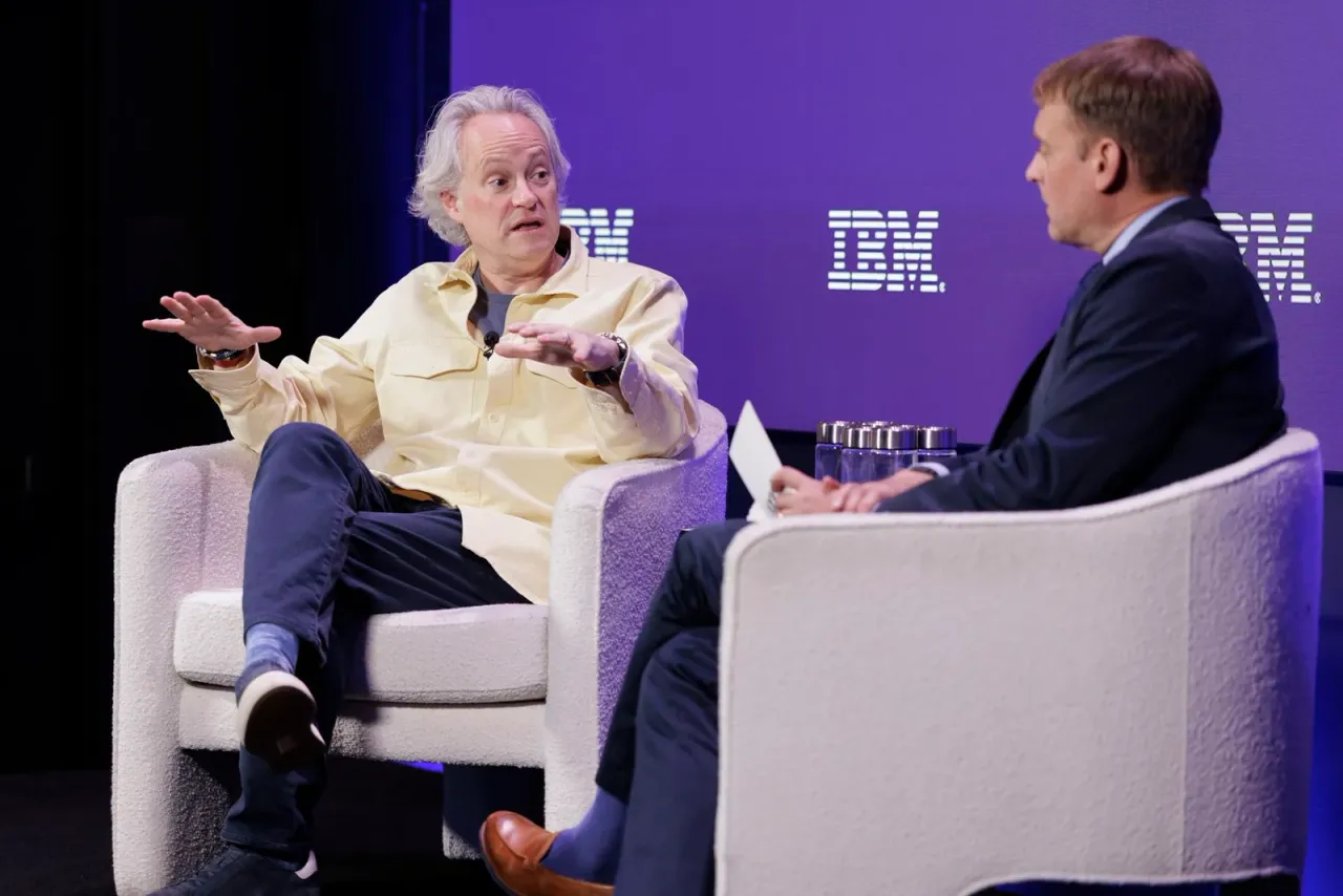 Two men sit in light armchairs on a stage with a purple IBM backdrop; the left man wears a pale yellow shirt and gestures, while the right man in a dark suit listens.