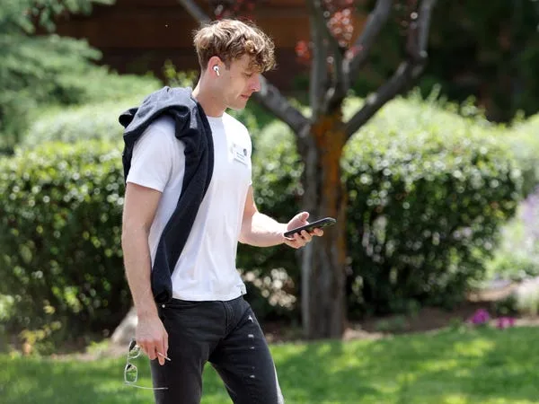 A man wearing earbuds walks outdoors while looking at a smartphone in a sunny garden setting.